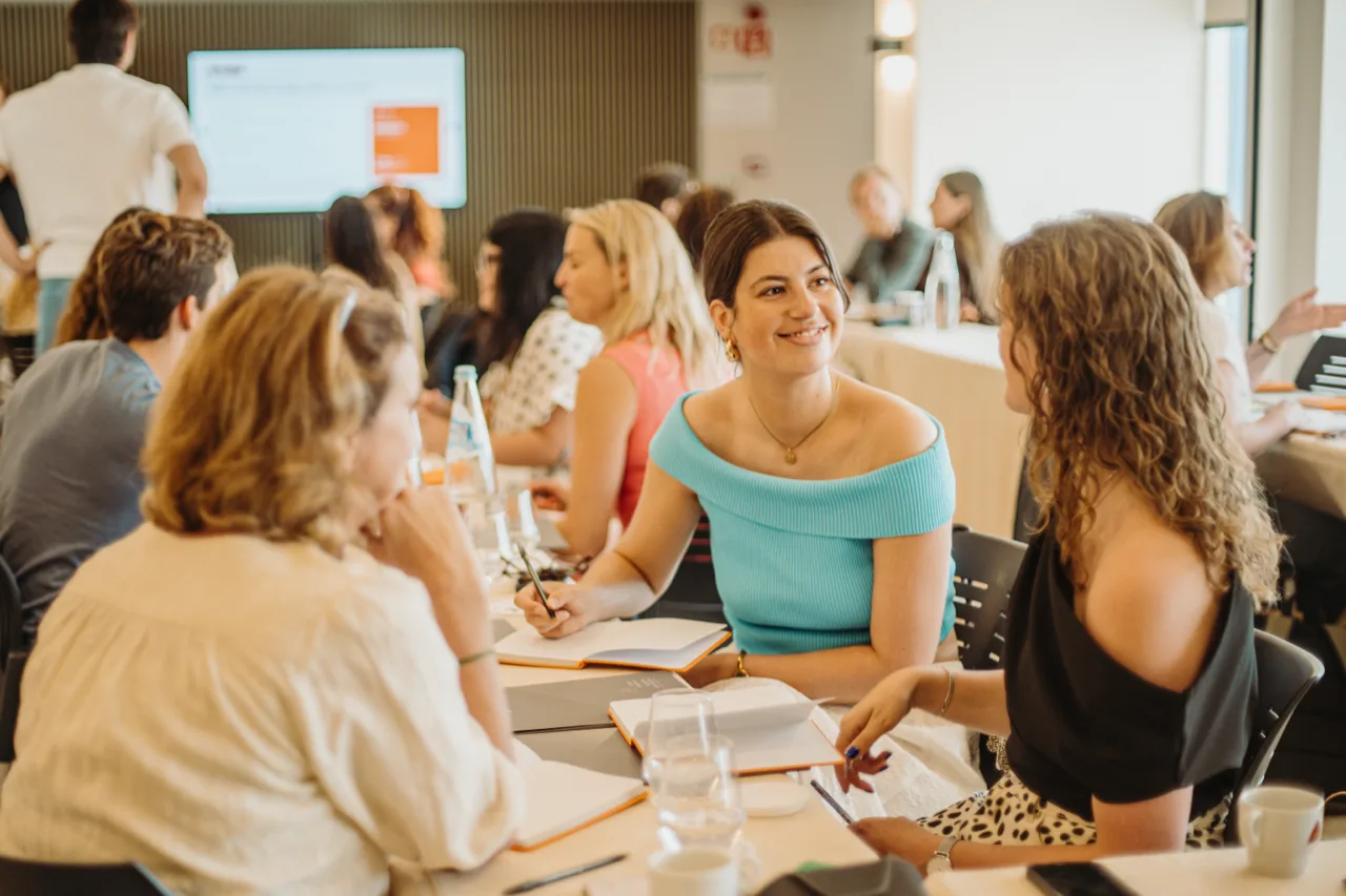 A woman smiling at a colleague across a table during a workshop, a full room of people behind them.